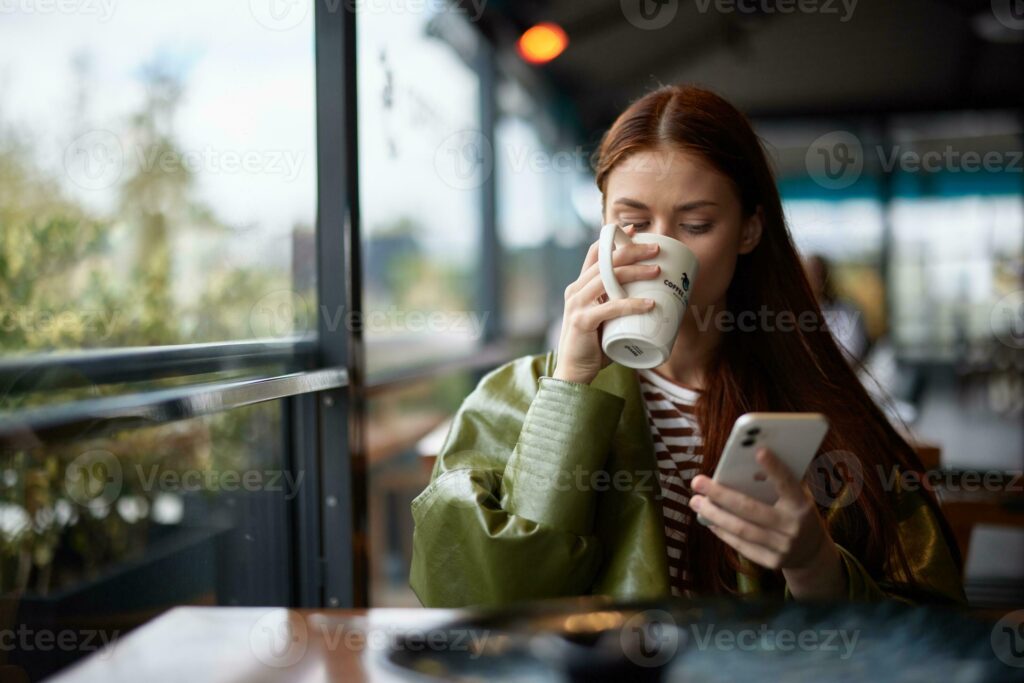 woman sitting in a cafe drinking coffee from a mug with phone in hand bloggergraphing food content for social media portrait of a stylish girl with red hair in the autumn photo